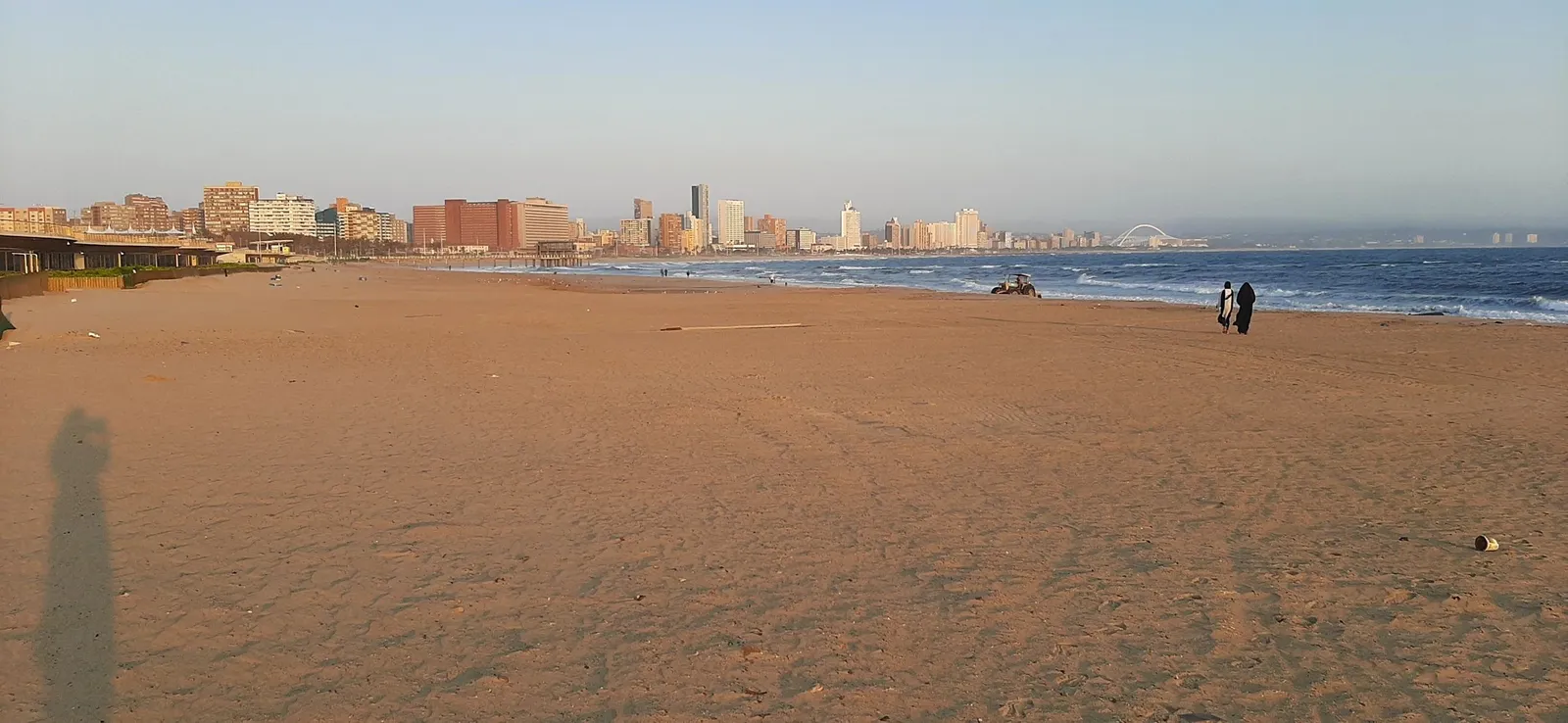 Spacious beach with city skyline and two people walking.