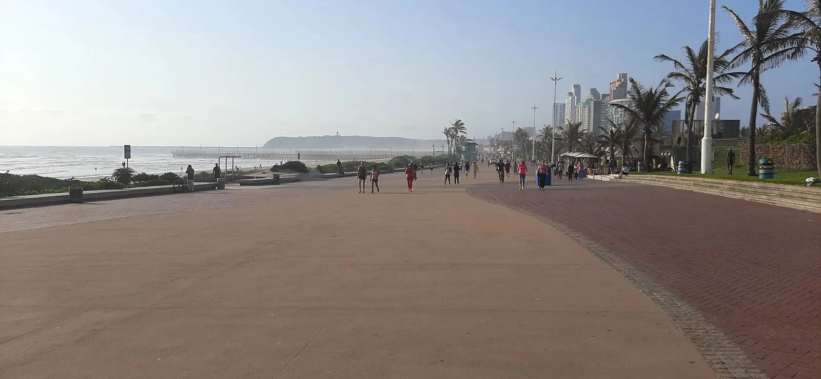 People walking along a beachfront promenade with city buildings in the background.